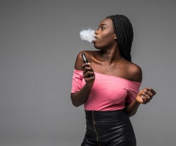 African woman smoking or vaping e-cig or electronic cigarette holding a mod with a lot of clouds isolated on black background